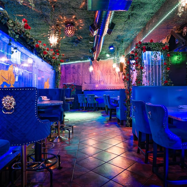 Babylon Cafe's interior with blue velvet seating, rose garlands, and Middle Eastern-inspired decor under colorful lighting.