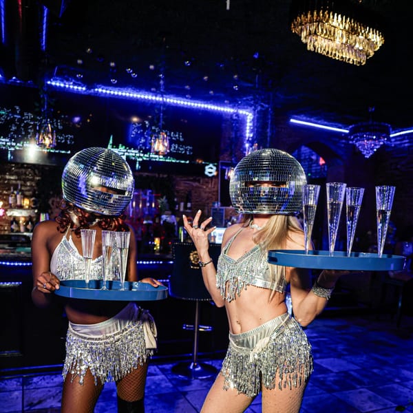 Two women in disco ball helmets and sparkly outfits hold trays of champagne flutes in a dark, blue-lit club.