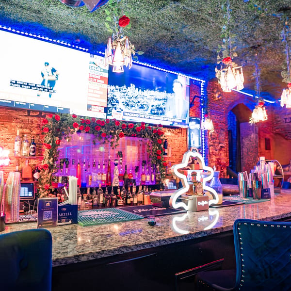 A vibrant bar at Babylon Cafe with bottles, red roses, blue lighting, and TVs showing sports.