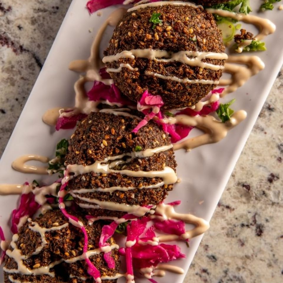 Three falafel balls drizzled with tahini sauce, served on a white plate with pickled red cabbage.