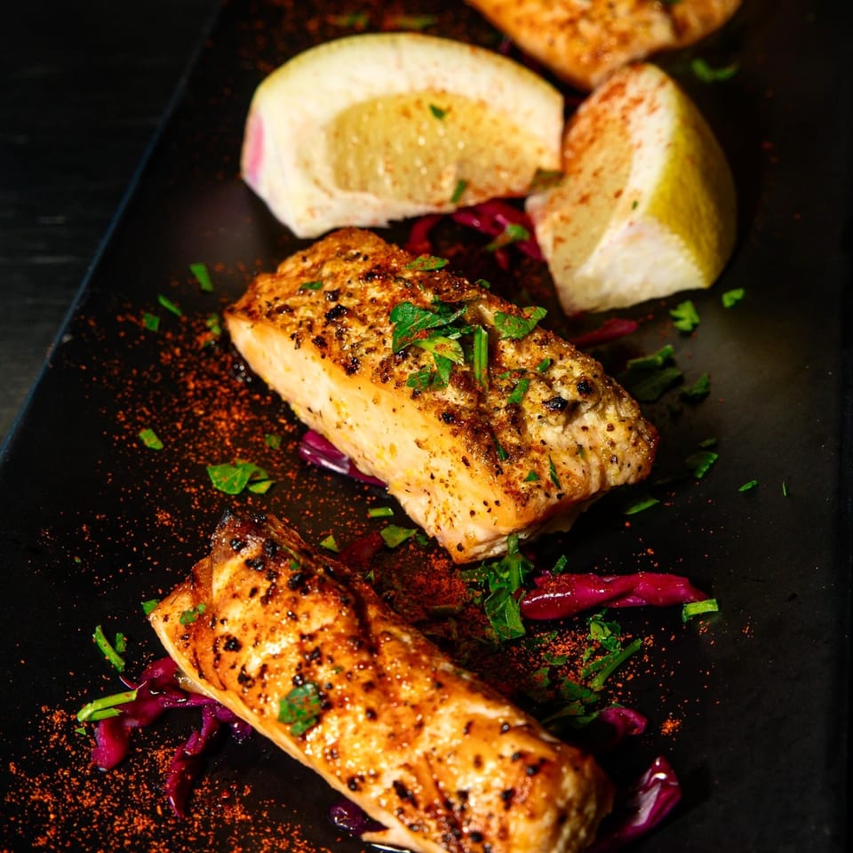 Two grilled salmon fillets with herbs, spices, pickled cabbage, and lemon wedges on a dark plate.