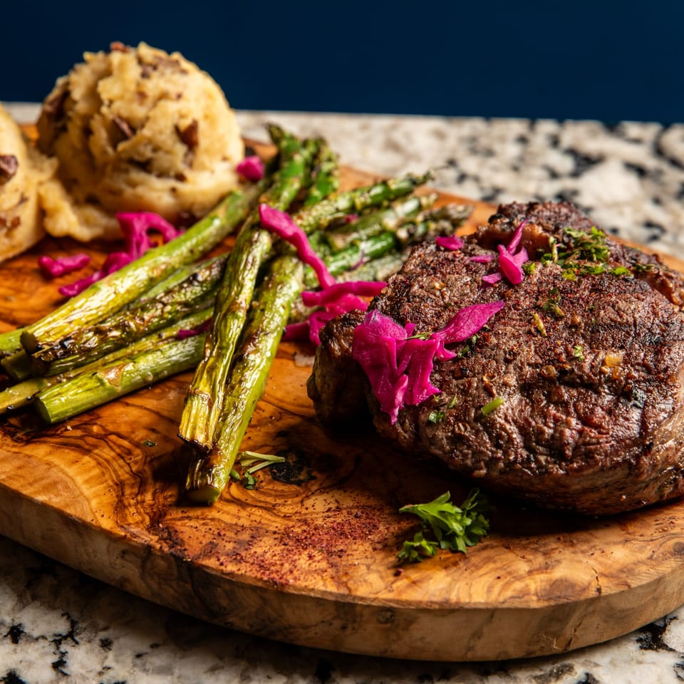 Grilled steak with asparagus, mashed potatoes, and pickled red onions on a wooden board.