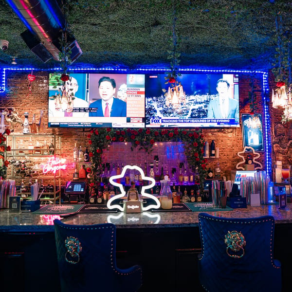 A vibrant bar at Babylon Cafe with neon lights, liquor bottles, and TVs displaying news.
