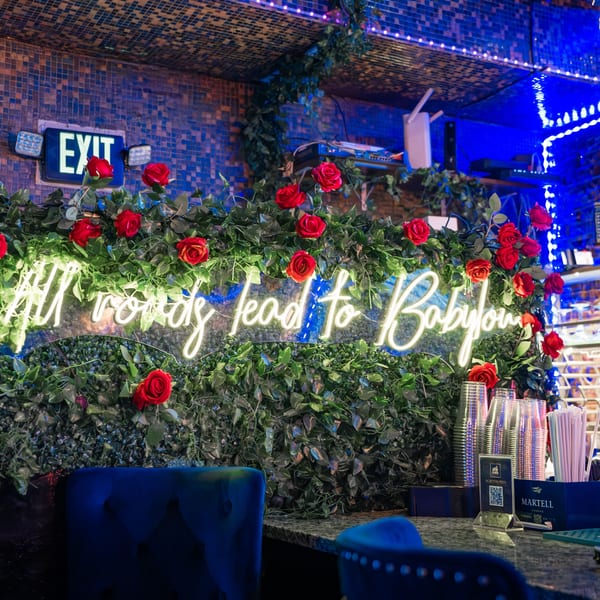 A neon sign reading "All roads lead to Babylon" on a green wall with red roses, above a bar.