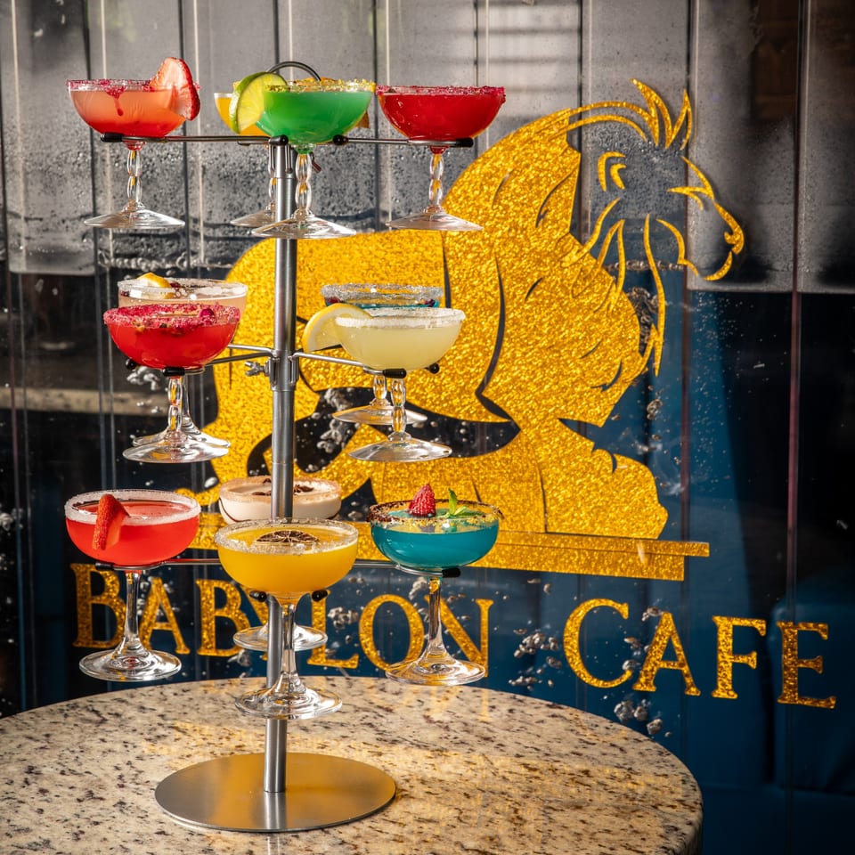 Colorful cocktails on a tiered stand, with the Babylon Cafe logo and horse emblem in the background.