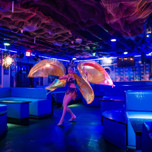 A belly dancer in a sequined costume with golden wings performs in a dark, blue-lit room.