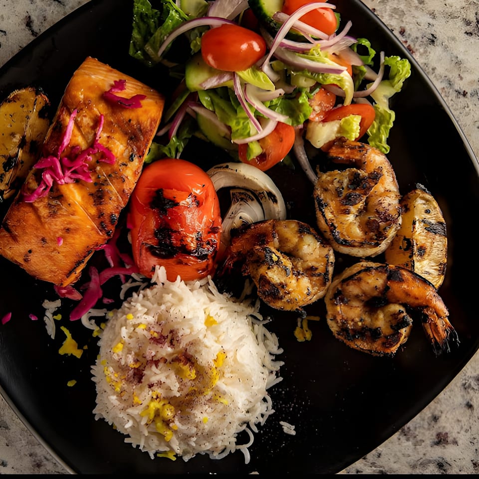 Grilled salmon, shrimp, tomato, onion, and lemon with rice and salad on a black plate.