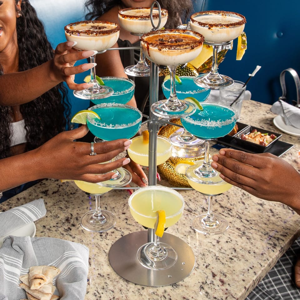 Hands reaching for a multi-tiered stand with various colorful cocktails at Babylon Cafe.