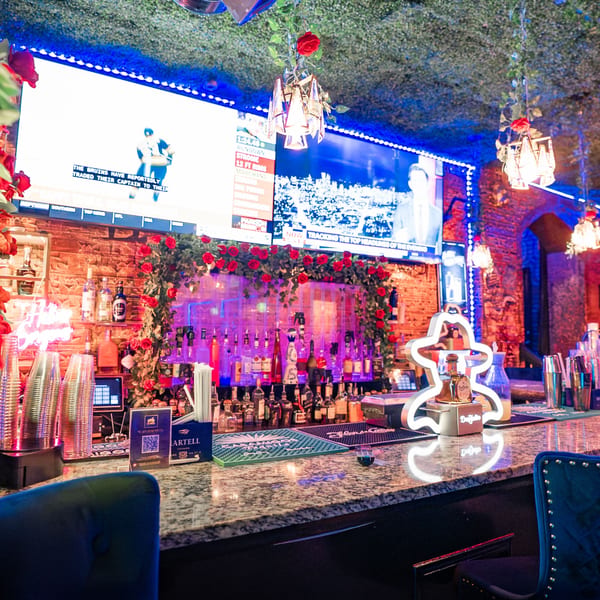 A vibrant bar at Babylon Cafe with neon lights, liquor bottles, and TVs showing sports news.