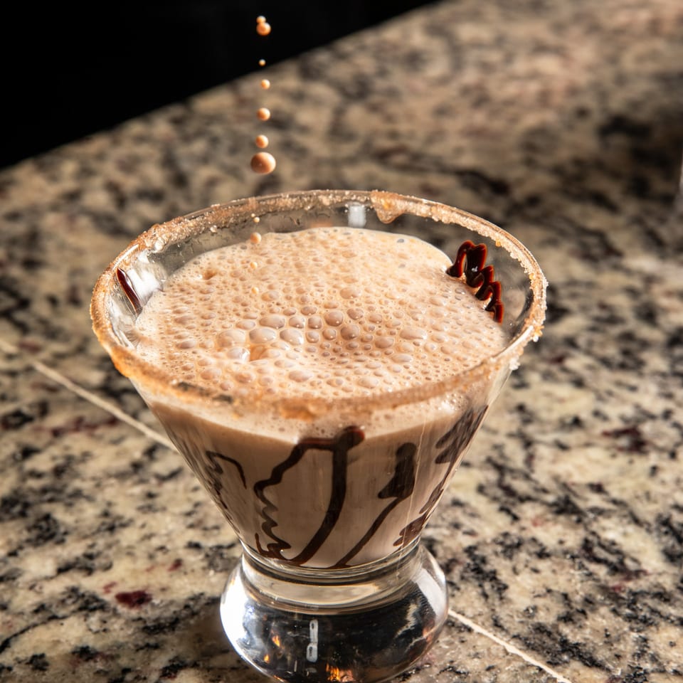 A frothy, light brown martini with chocolate drizzle and a sugar-rimmed glass, with liquid drops falling.