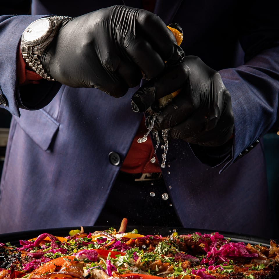 Gloved hands squeeze a lemon over a vibrant Middle Eastern dish with meat, red cabbage, and herbs.